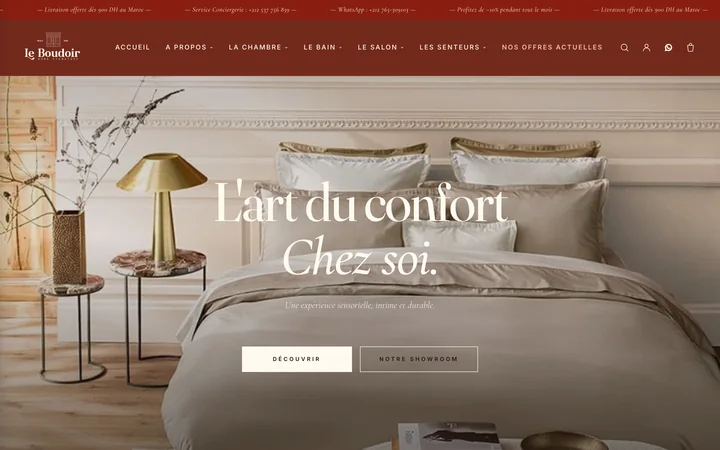 The Le Boudoir homepage on a desktop screen: a terracotta announcement bar, the Le Boudoir wordmark with a French navigation, and a photograph of a made bed in beige linen with a brass lamp and dried flowers, carrying the headline "L’art du confort chez soi." above two outlined buttons.