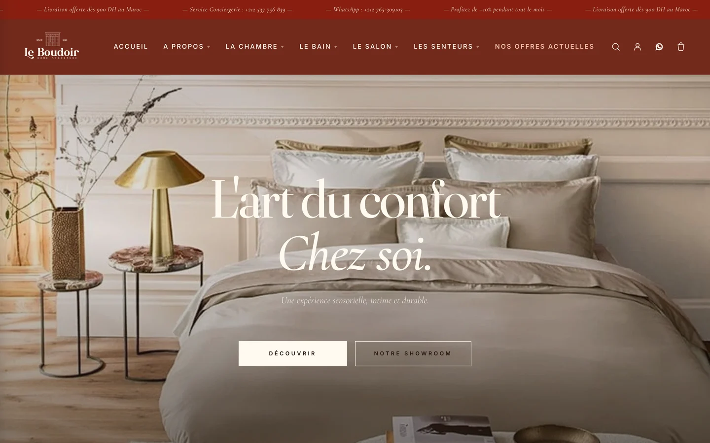The Le Boudoir homepage on a desktop screen: a terracotta announcement bar, the Le Boudoir wordmark with a French navigation, and a photograph of a made bed in beige linen with a brass lamp and dried flowers, carrying the headline "L’art du confort chez soi." above two outlined buttons.